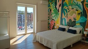 A bedroom with a bed, a colorful tropical mural, a radiator, and a view of the city through a sliding door.