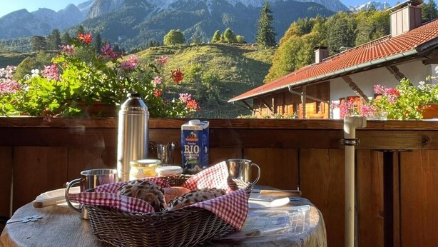 A wooden table with a basket, a thermos, a milk carton, and a metal mug on a balcony with a mountain view. - Apartment 'Höllental' with Mountain View, Balcony, and Wi-Fi (Grainau)