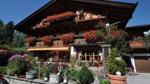 A traditional wooden chalet with balconies adorned with flowers, a stone wall, and a garden with potted plants. - Apartment 'Höllental' with Mountain View, Balcony, and Wi-Fi (Grainau)