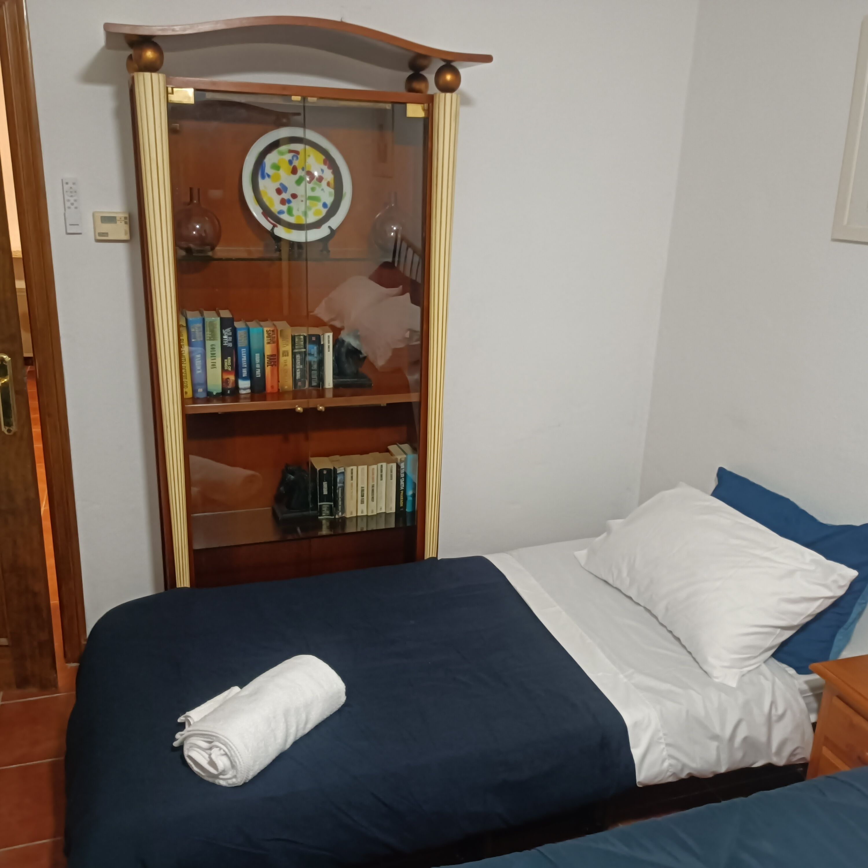 Un dormitorio con cama, almohadas, una mesita de noche y un armario de madera con libros.