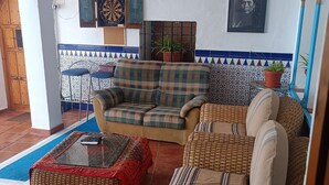 Un salon avec un canapé, une table basse et un fauteuil en osier. Il y a un jeu de fléchettes accroché au mur et un tableau encadré au-dessus du canapé.