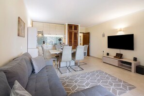Una sala de estar moderna con un sofá gris, un comedor con mesa y sillas, una cocina al fondo y un televisor montado en la pared.