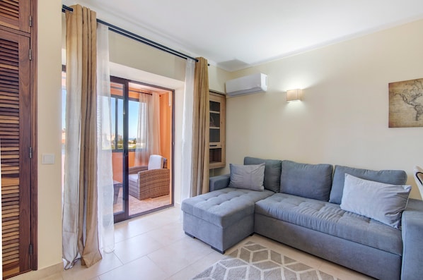 A living room with a grey sectional sofa, a glass door leading to an outdoor area, an air conditioning unit, and a wall-mounted light fixture.
