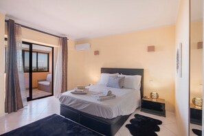 Un dormitorio moderno con una cama, una mesita de noche pequeña, una alfombra negra y una puerta de cristal corredera que da a un balcón.