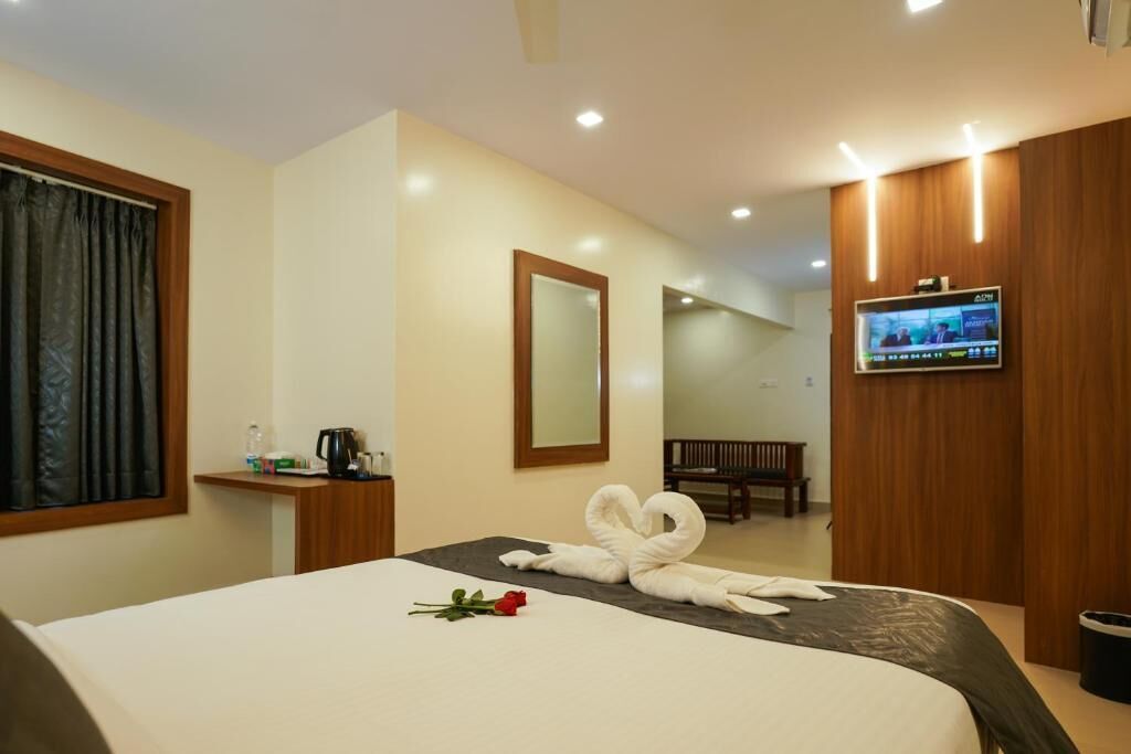 A hotel room with a bed, a TV mounted on the wall, a wooden bench, and a small table with a kettle and cups.