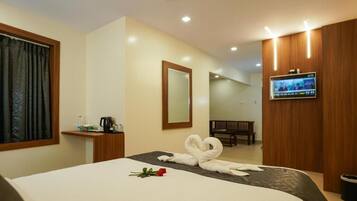A hotel room with a bed, a TV mounted on the wall, a wooden bench, and a small table with a kettle and cups.