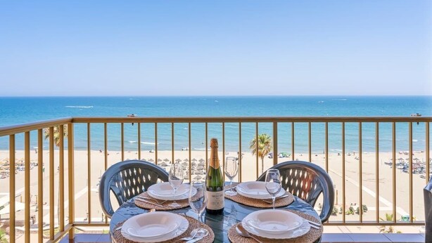 Une table dressée pour deux personnes, avec une bouteille de champagne, donnant sur une plage bordée de palmiers et parsemée de parasols.