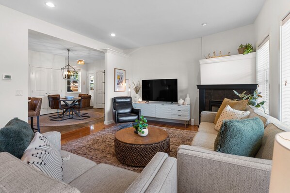 A modern living room with a sofa, coffee table, and TV. There is a fireplace and a dining area in the background.