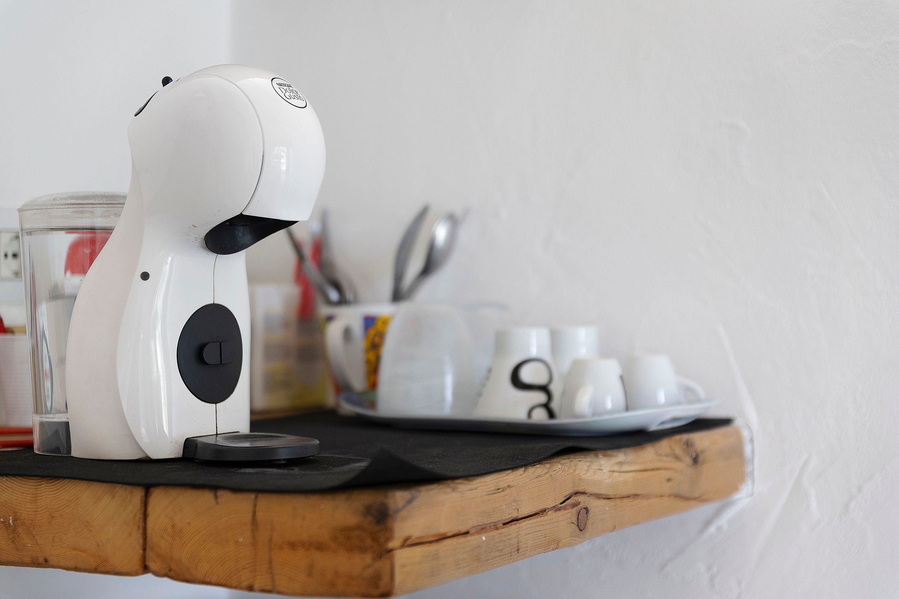 A white coffee maker on a wooden shelf with a glass of water, a tray with cups, and a spoon.