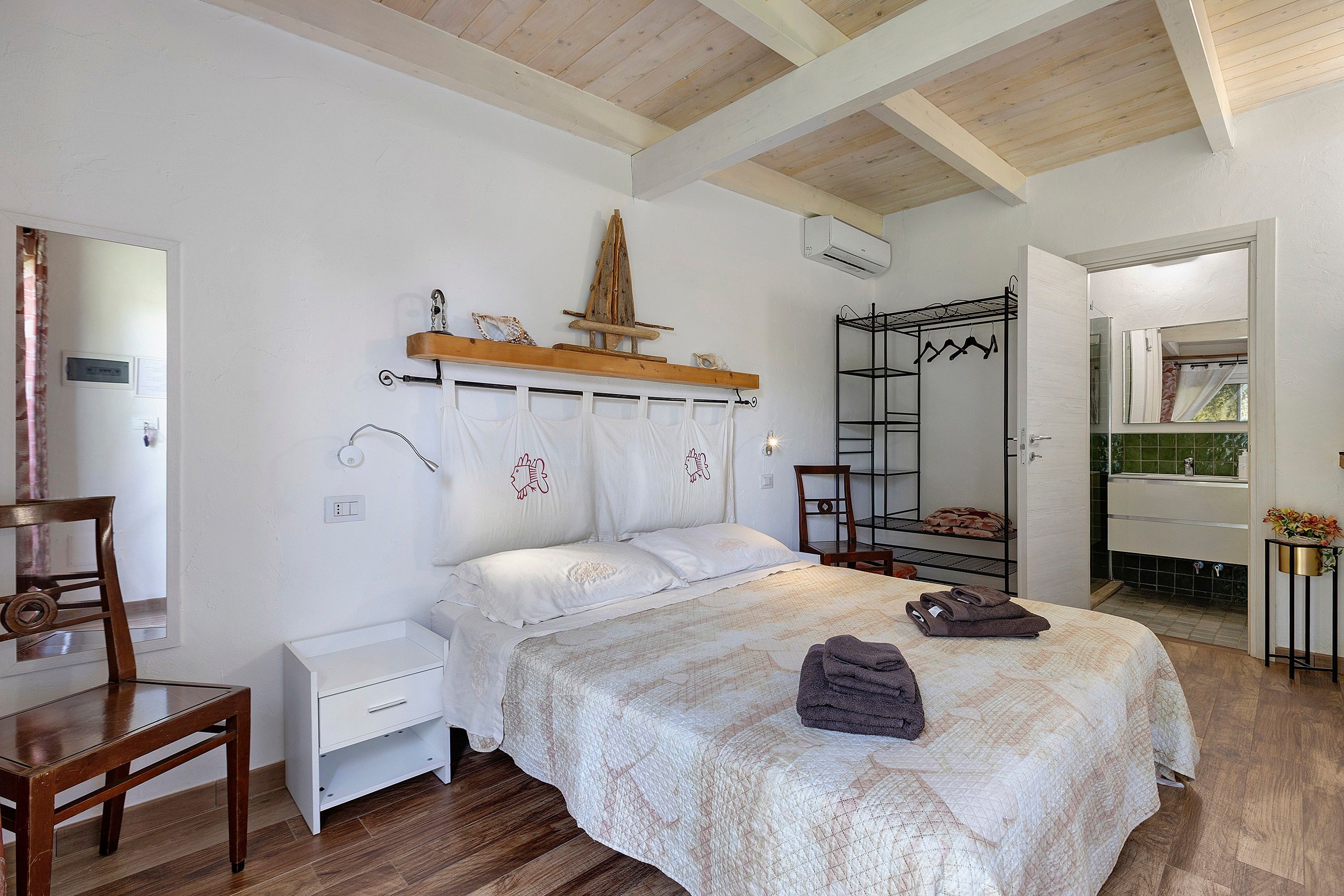 A bedroom with a bed, a chair, a bedside table, a wooden shelf with decorative items, and a wardrobe with a mirror.