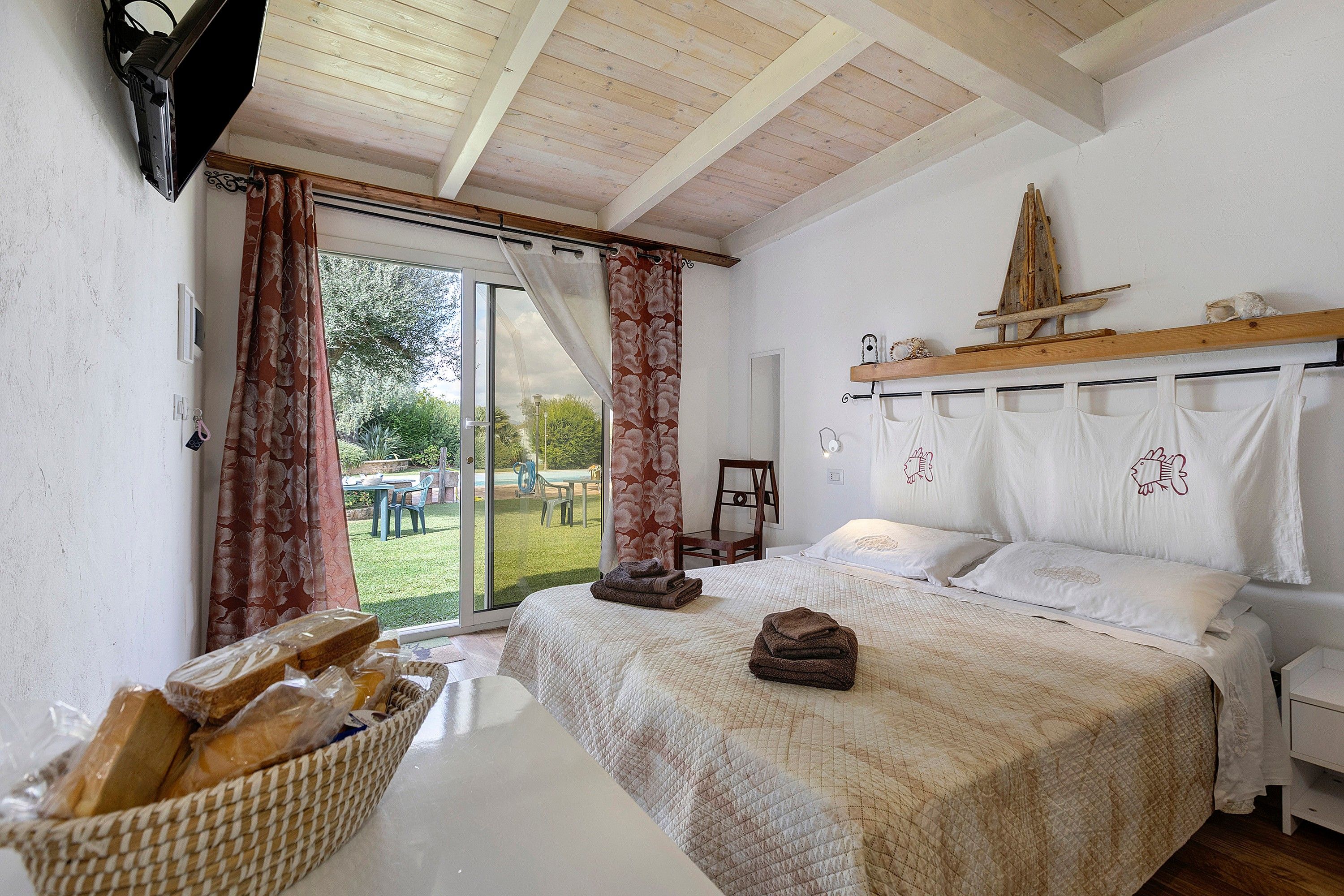A bedroom with a large bed, a wooden headboard, a basket of bread, and a view of the outdoors.