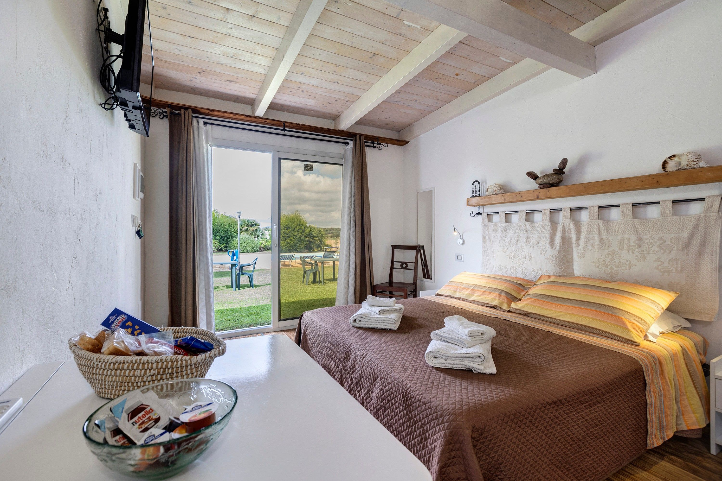 A bedroom with a bed, a table with a bowl of snacks, a chair, and a view of the outdoors through a sliding door.