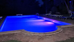 A swimming pool with a blue-lit edge, surrounded by a grassy area and trees.