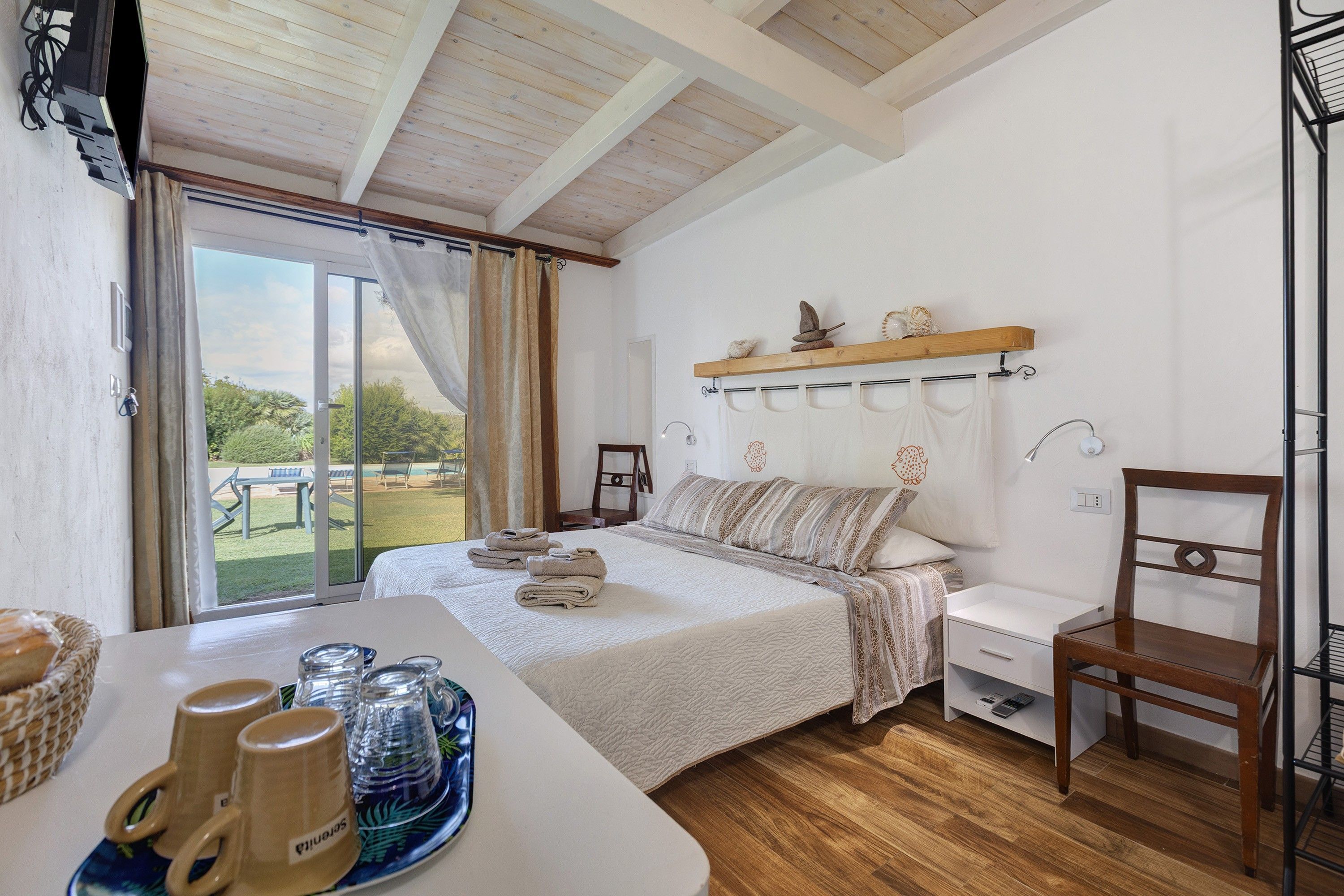 A bedroom with a bed, a bedside table with towels, a chair, a shelf with decorative items, and a view of the outdoors.