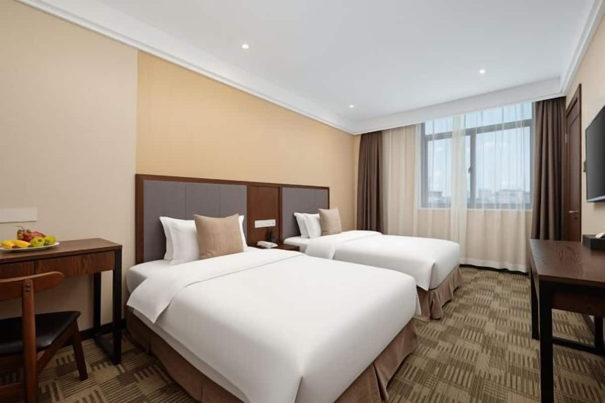 A hotel room with two beds, a desk, a chair, a TV, and a large window with curtains.