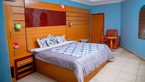 A bedroom with a wooden bed, a nightstand, a chair, and a small table. - The Kings Hotel and Suites  (Ibadan)
