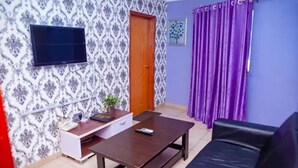 A room with a patterned wallpaper, a television mounted on the wall, a wooden table, a black sofa, and a purple curtain. - The Kings Hotel and Suites  (Ibadan)