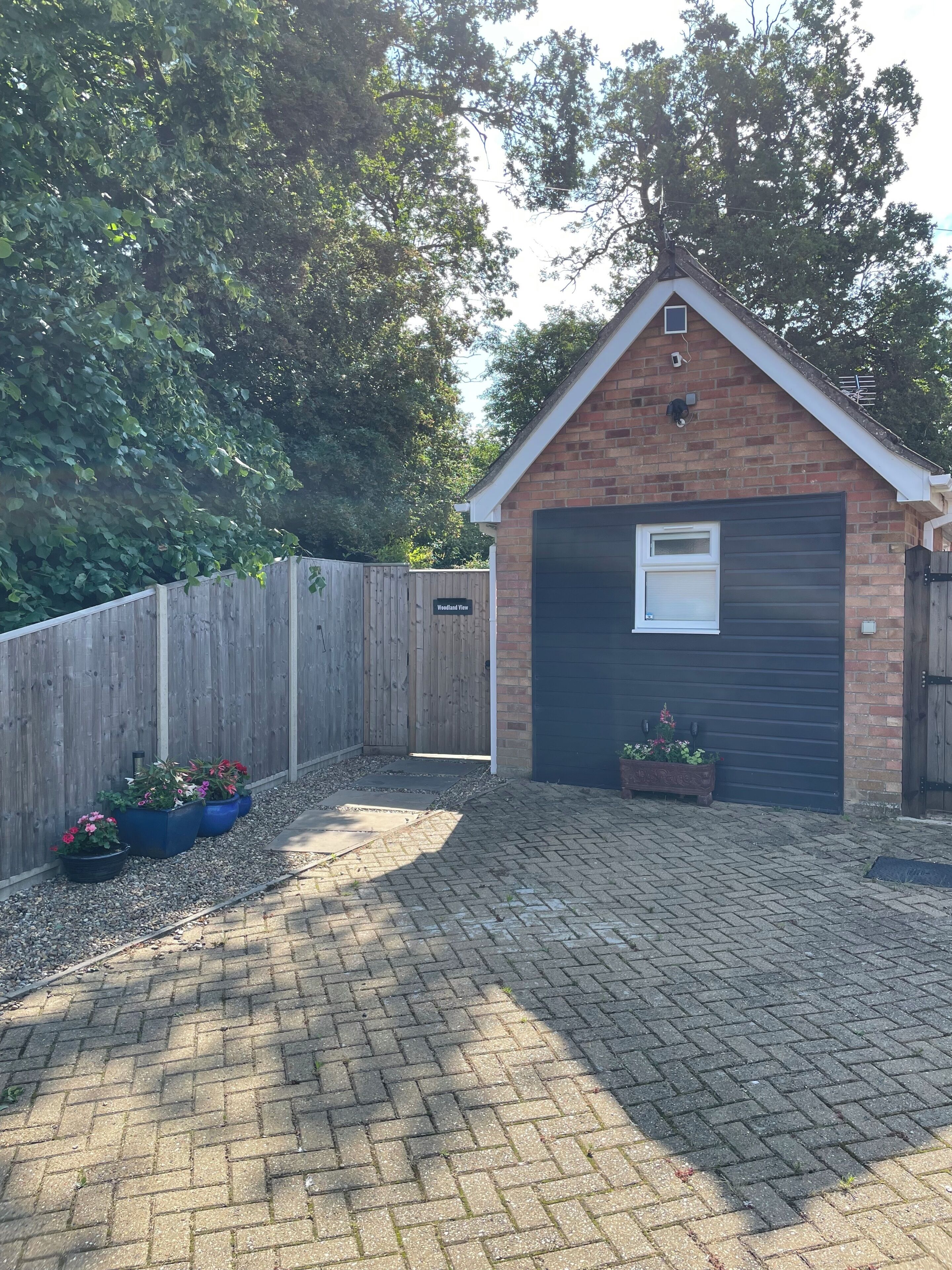 Un garage en briques avec une porte sombre, une petite fenêtre et une clôture en bois à l’arrière-plan.
