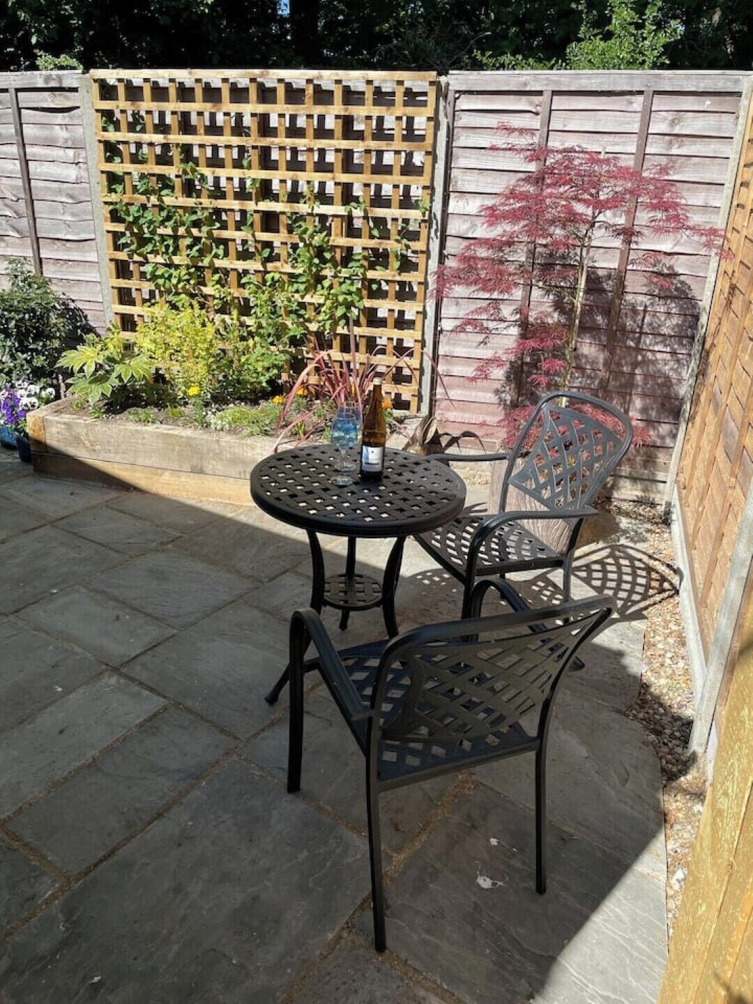 An outdoor patio with a round table and two chairs, a bottle of wine, and a wooden lattice fence with plants behind it.