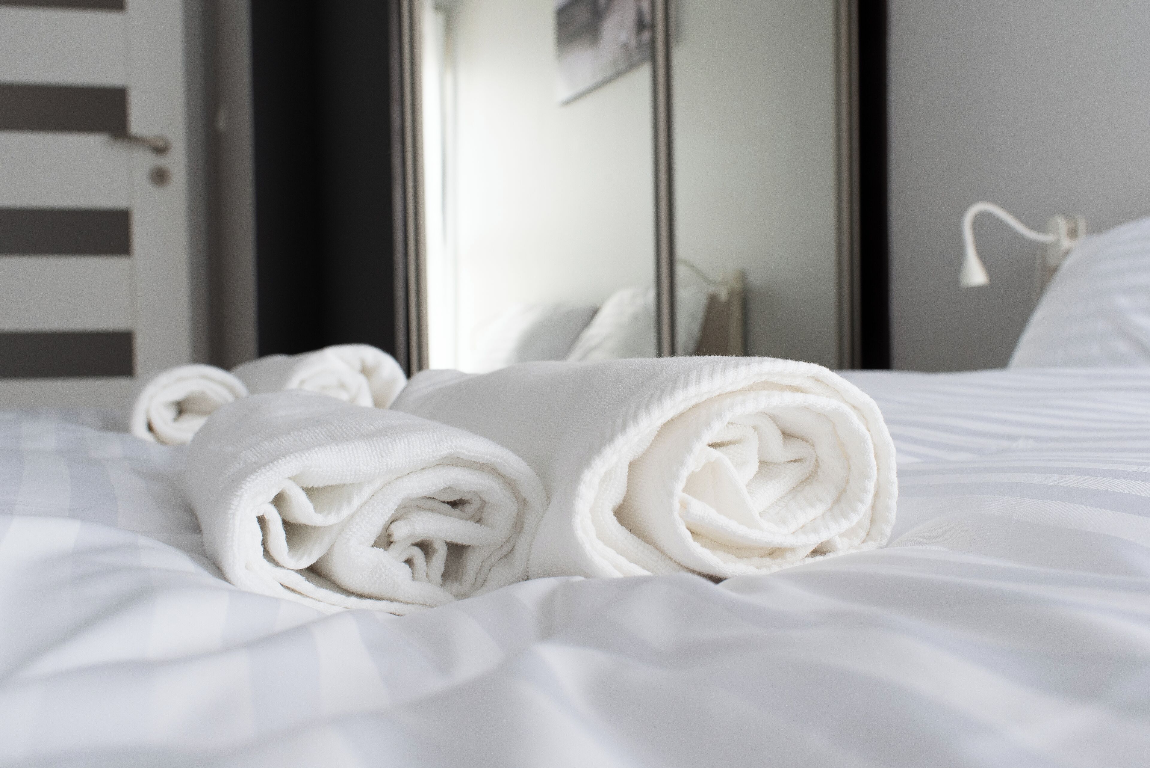 A neatly made bed with rolled white towels and a mirror reflecting the room's interior.