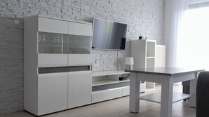 A modern living room with a white brick wall, a flat-screen TV mounted above a white cabinet, and a dining table with a lamp. - RentPlanet - Apartament przy Lotnisku (Poznań)