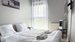 A neatly made bed with white linens and rolled towels, a bedside table with a lamp, a radiator, and a wall-mounted TV. - RentPlanet - Apartament przy Lotnisku (Poznań)