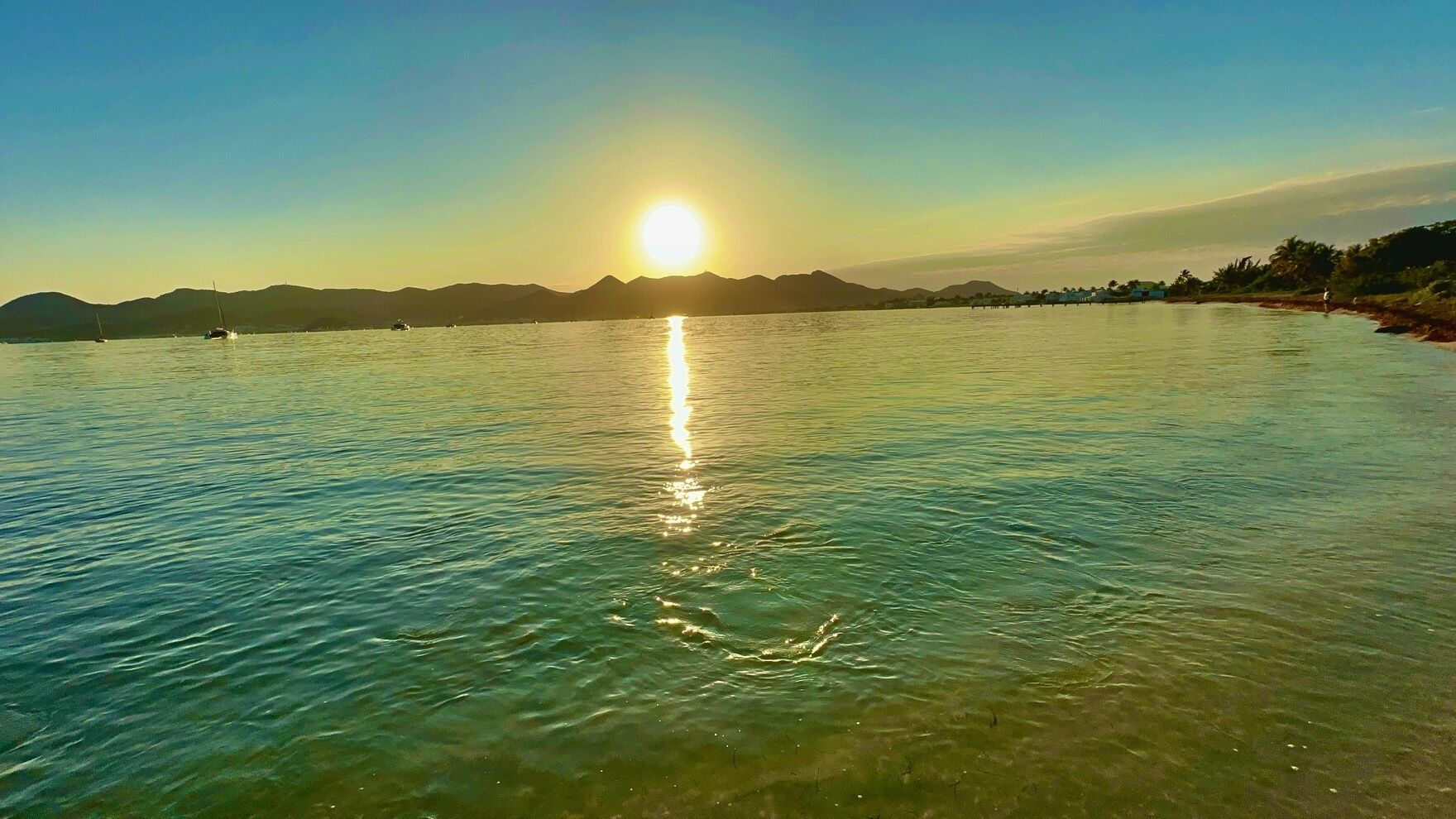 Une étendue d'eau calme où se reflète le soleil, avec des montagnes lointaines et un ciel dégagé.
