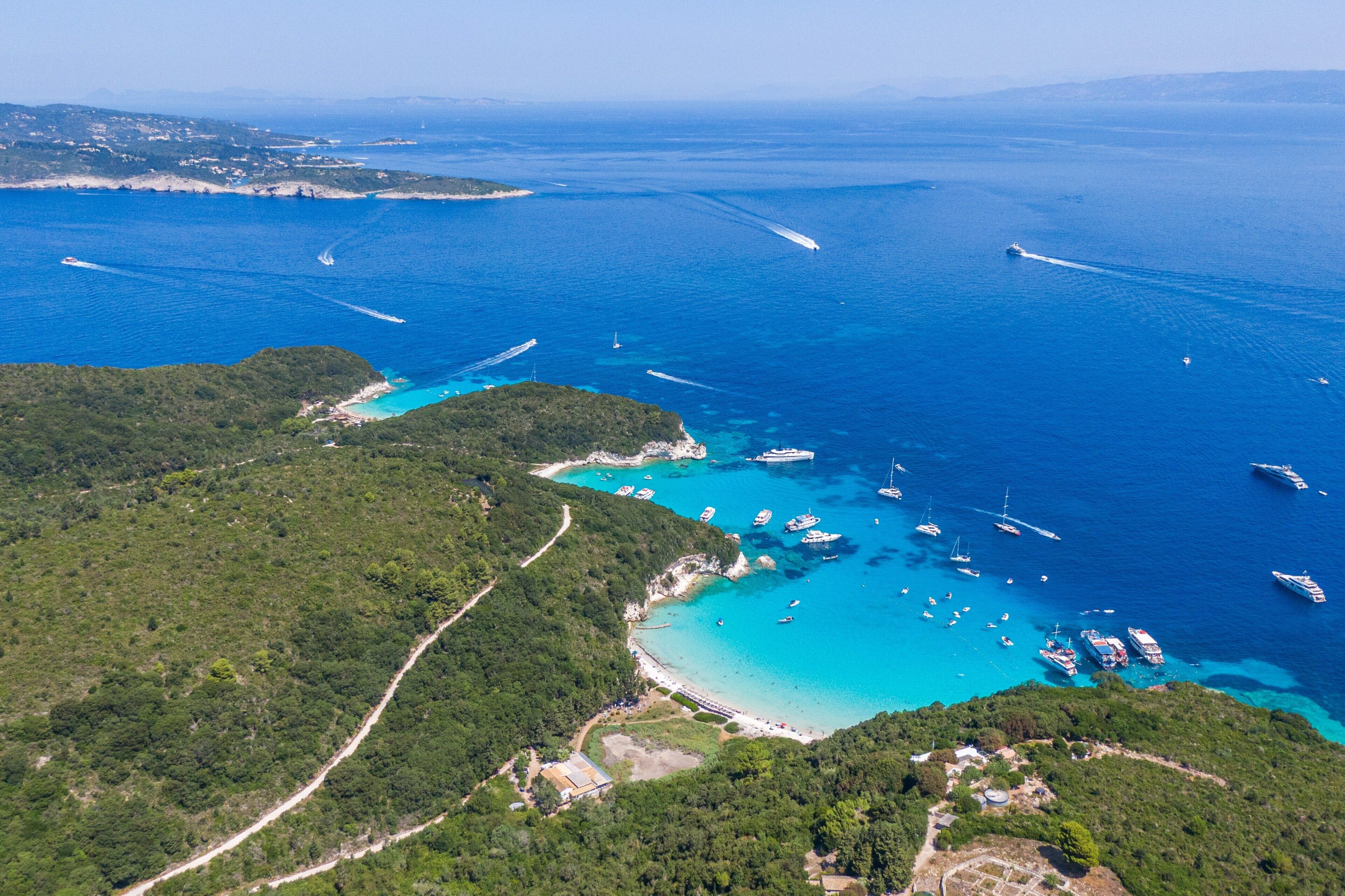 Eine Vogelperspektive zeigt eine Küstenlandschaft mit klarem türkisblauem Wasser, Booten und einer sich durch die Landschaft schlängelnden Straße.