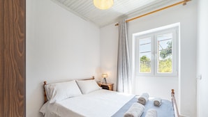 A bedroom with a bed, a bedside lamp, a window with curtains, and a view of greenery outside. - Apartment 'Ambelaki' with Sea View, Private Garden and Wi-Fi (Antípaxos)