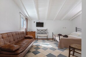 A room with a brown leather sofa, a wooden chair, a bed with a white bedspread, and a television mounted on the wall.