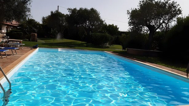 A rectangular swimming pool with clear blue water, surrounded by a stone deck and a grassy area with trees. - Studio 'Ulivo' with Shared Pool, Wi-Fi and Air Conditioning (Alghero)