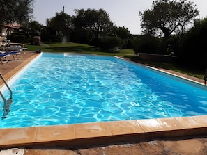 Une piscine rectangulaire à l’eau d’un bleu cristallin, entourée d’une terrasse en pierre et d’une zone herbeuse agrémentée d’arbres.