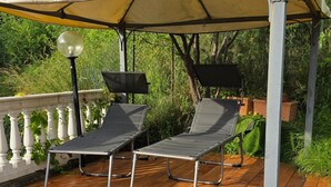 A covered outdoor seating area with two lounge chairs and a table, surrounded by greenery.