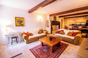 A living room with two sofas, a coffee table, a desk with a chair, a bookshelf, and a kitchen area with stone walls. - The Logis de la Bastide (Lauzerte)