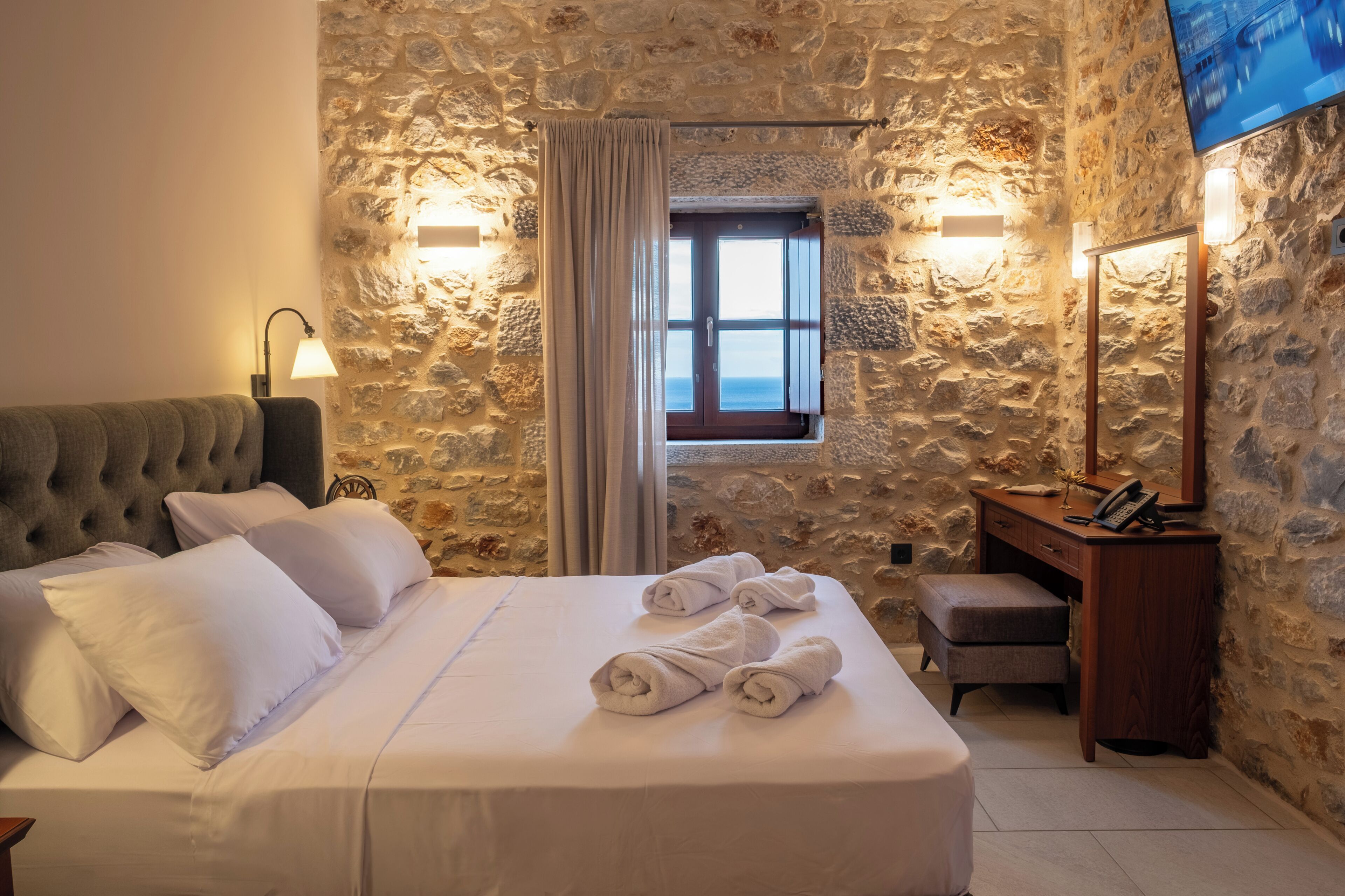 Un dormitorio con pared de piedra, cama con ropa de cama blanca, mesita de noche de madera, escritorio y televisor montado en la pared.