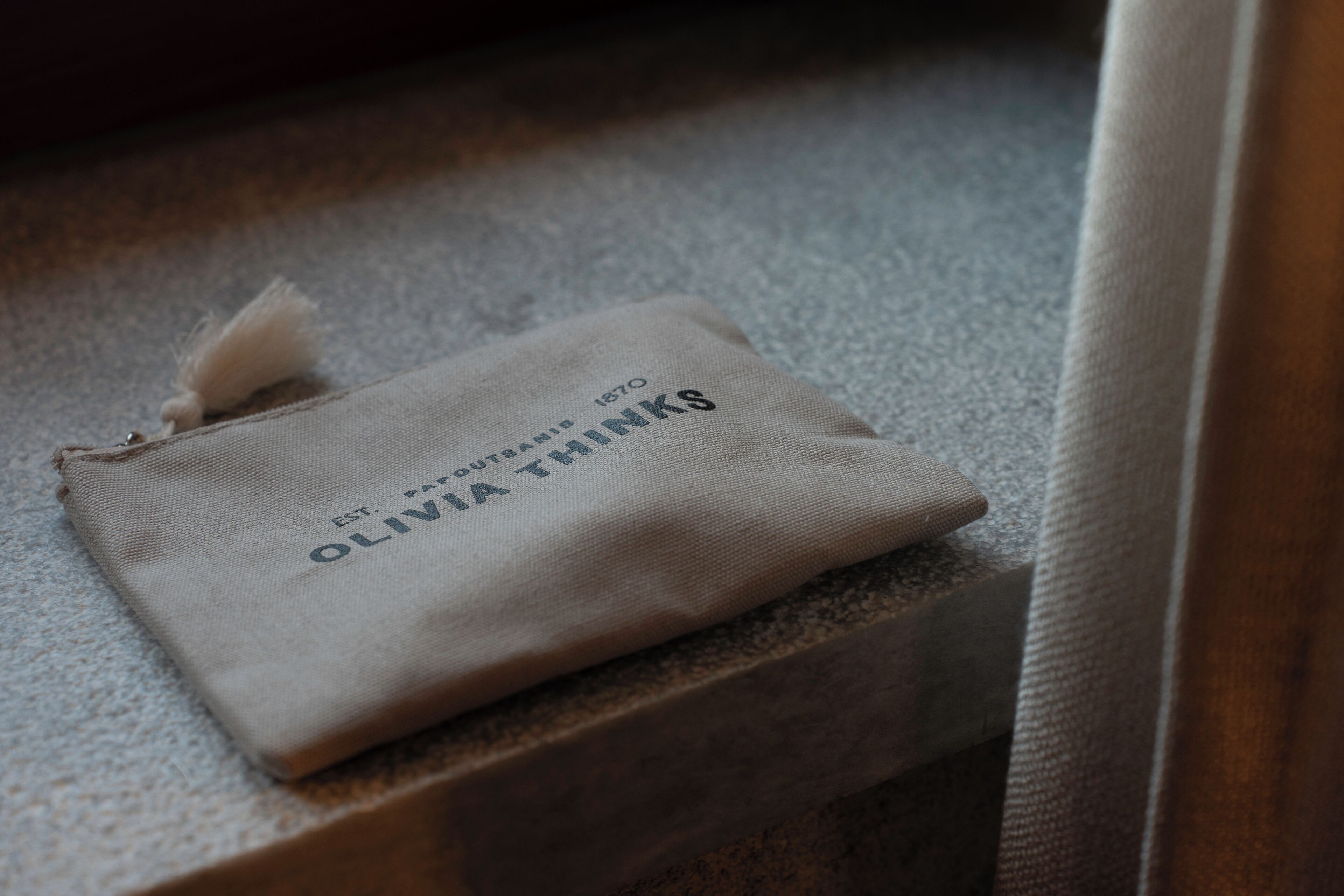 A small, beige fabric pouch with the text "OLIVIA THINKS" and "ESTD 1870" on a textured surface.