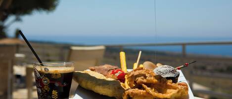 A meal consisting of fried fish, fries, a drink, and a dessert on a table with a view of the sea.