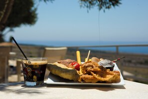 A meal consisting of fried fish, fries, a drink, and a dessert on a table with a view of the sea. - Anemos Suites Mani (East Mani)