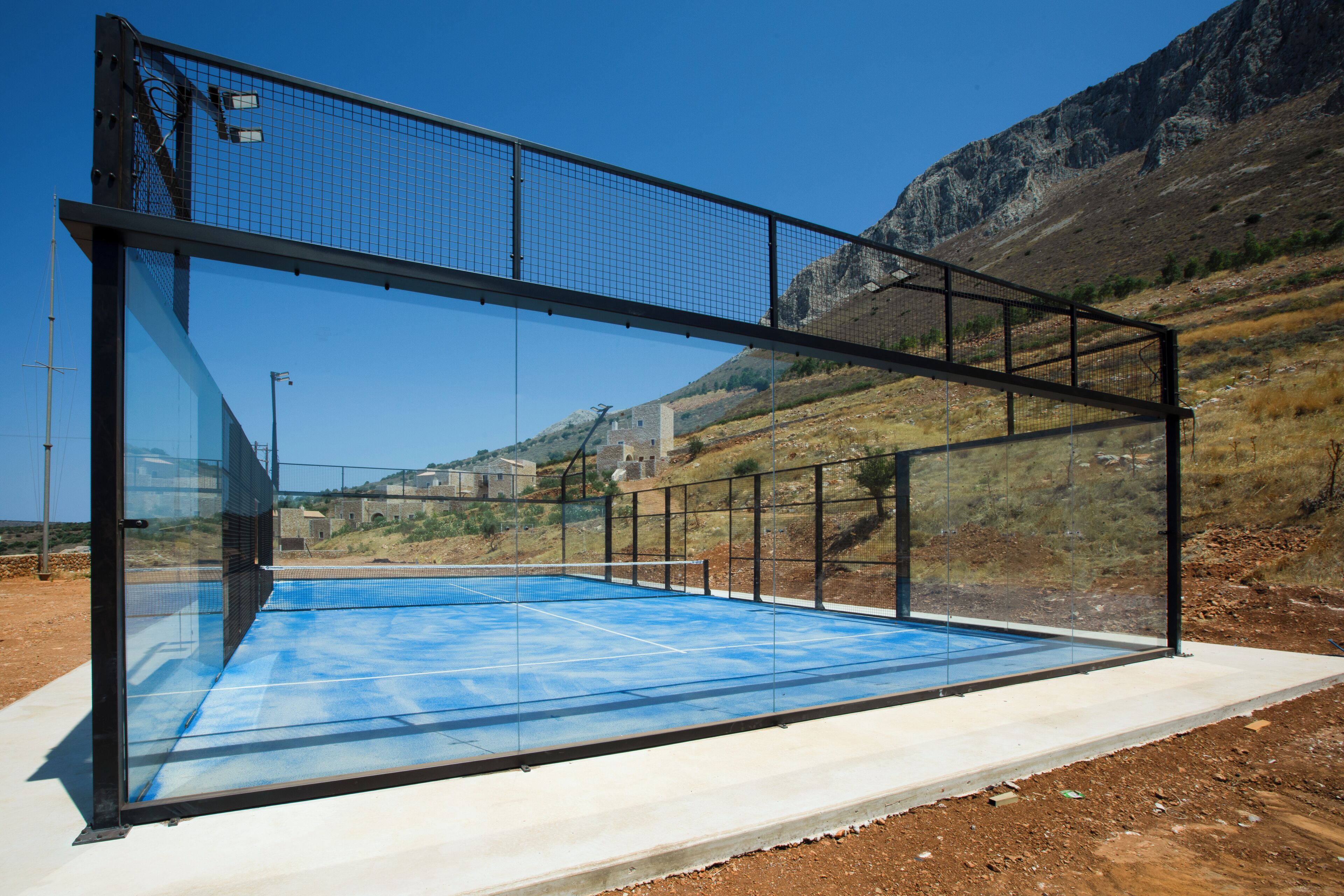 A modern outdoor tennis court with a glass enclosure, surrounded by a hilly landscape.