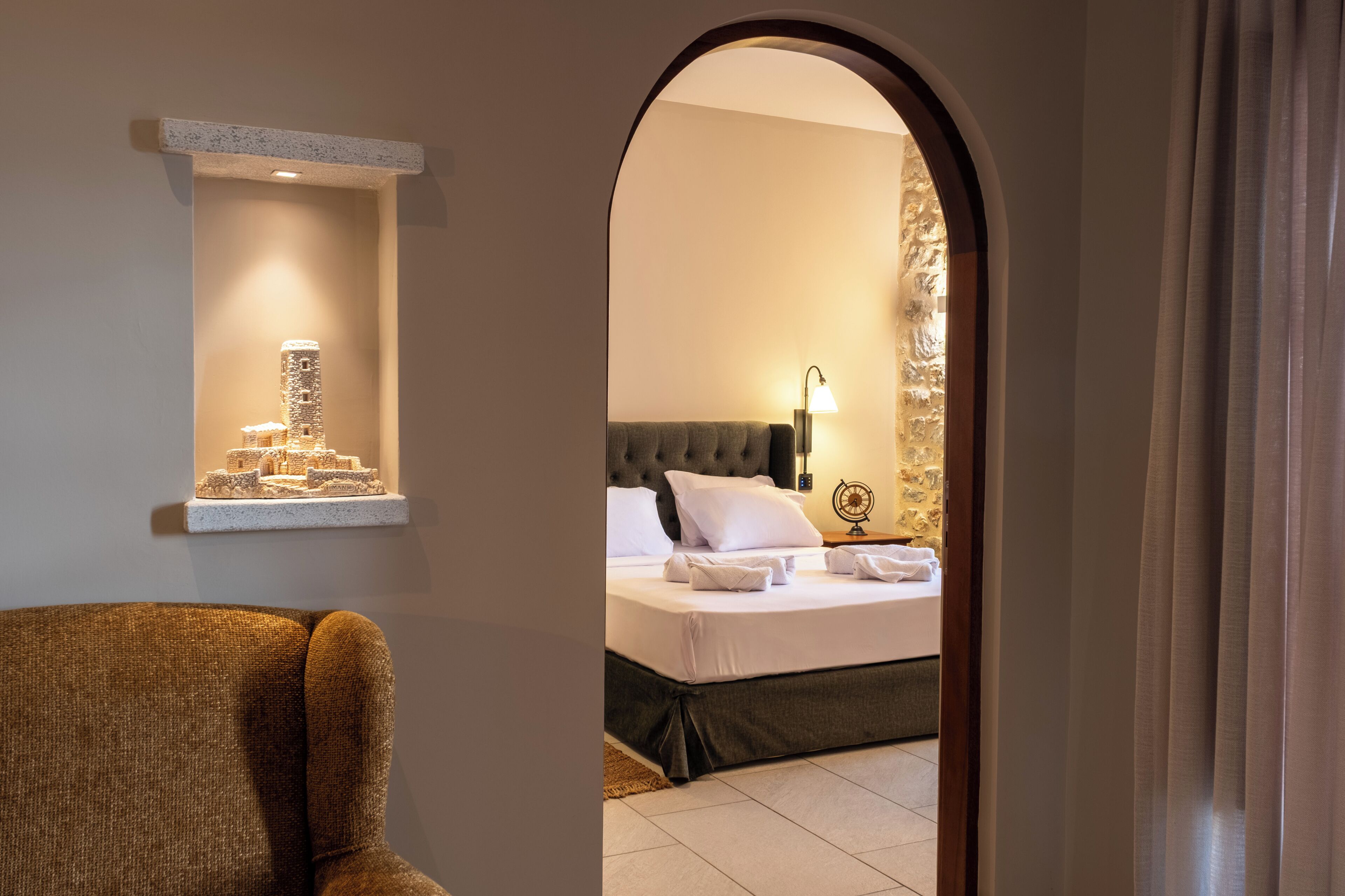 Ein Hotelzimmer mit Bett, Sessel, einer Wandablage mit Dekoration und Blick durch eine Bogenöffnung.