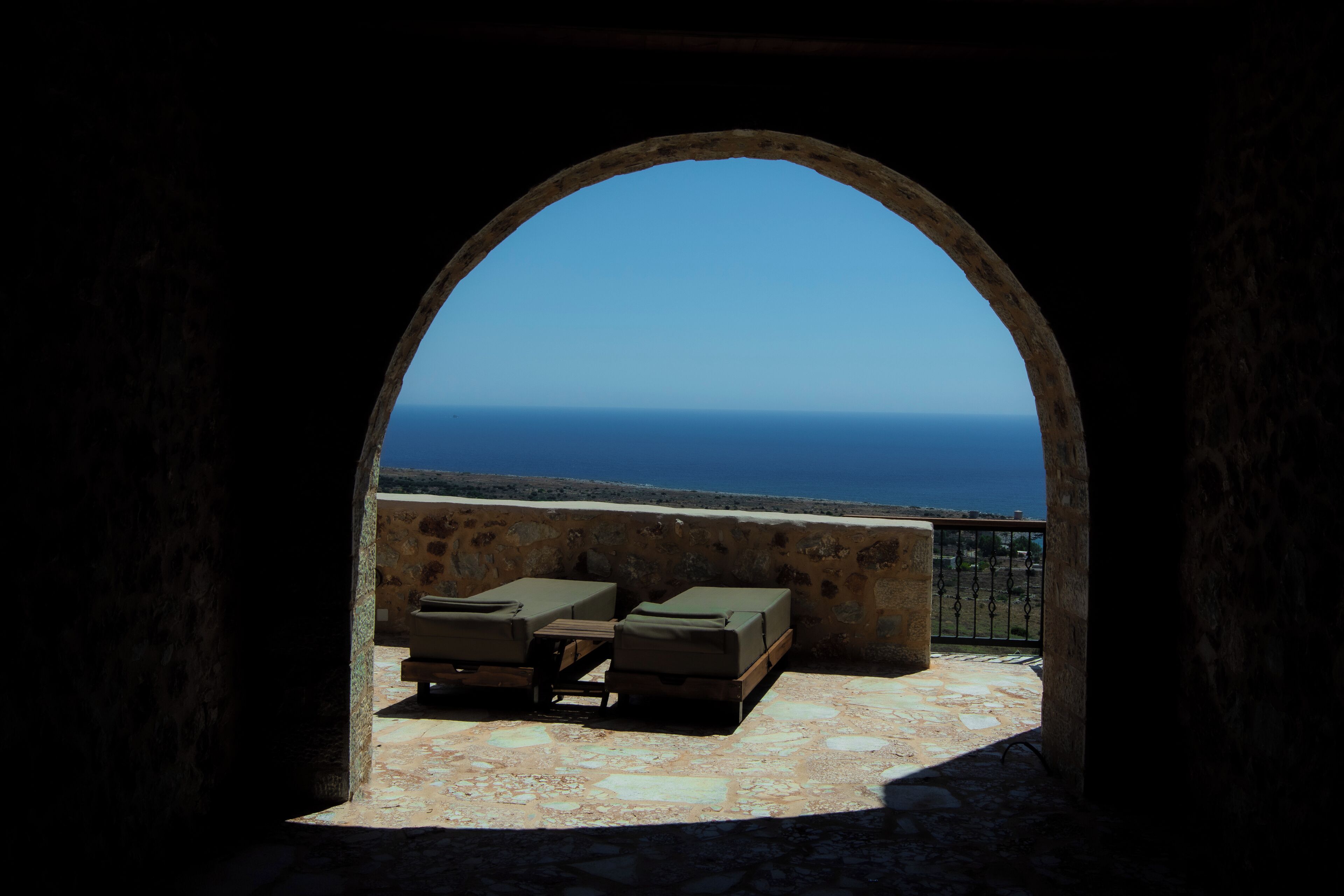 An arched outdoor seating area with stone walls and a view of the sea.
