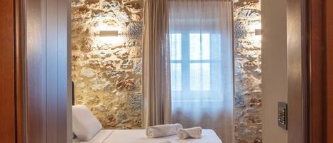 A bedroom with a stone wall, a wooden ceiling, a bed with white linens, and a window with curtains.
