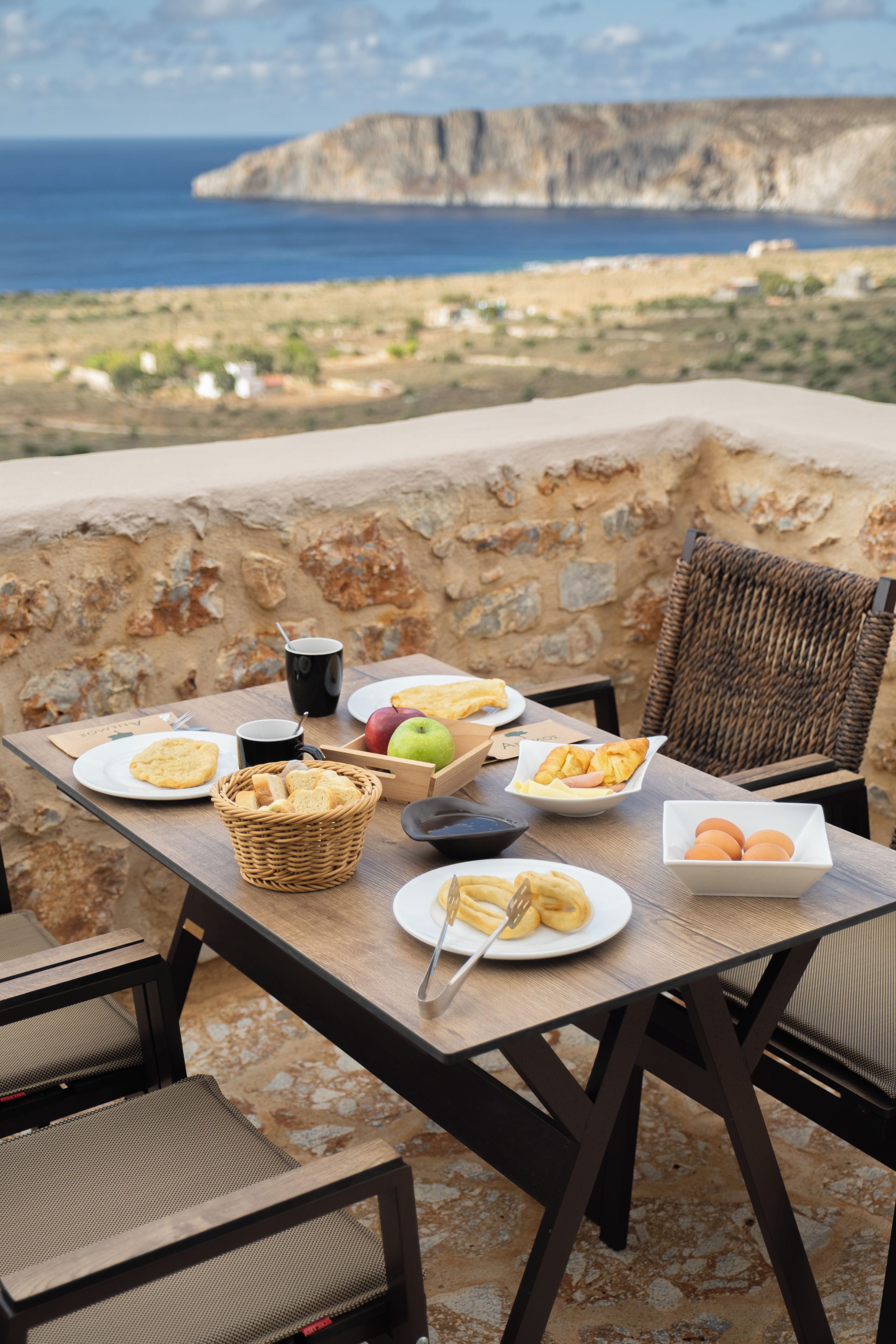 An outdoor dining area with a table set for two, featuring a variety of food items and a view of the sea and cliffs.