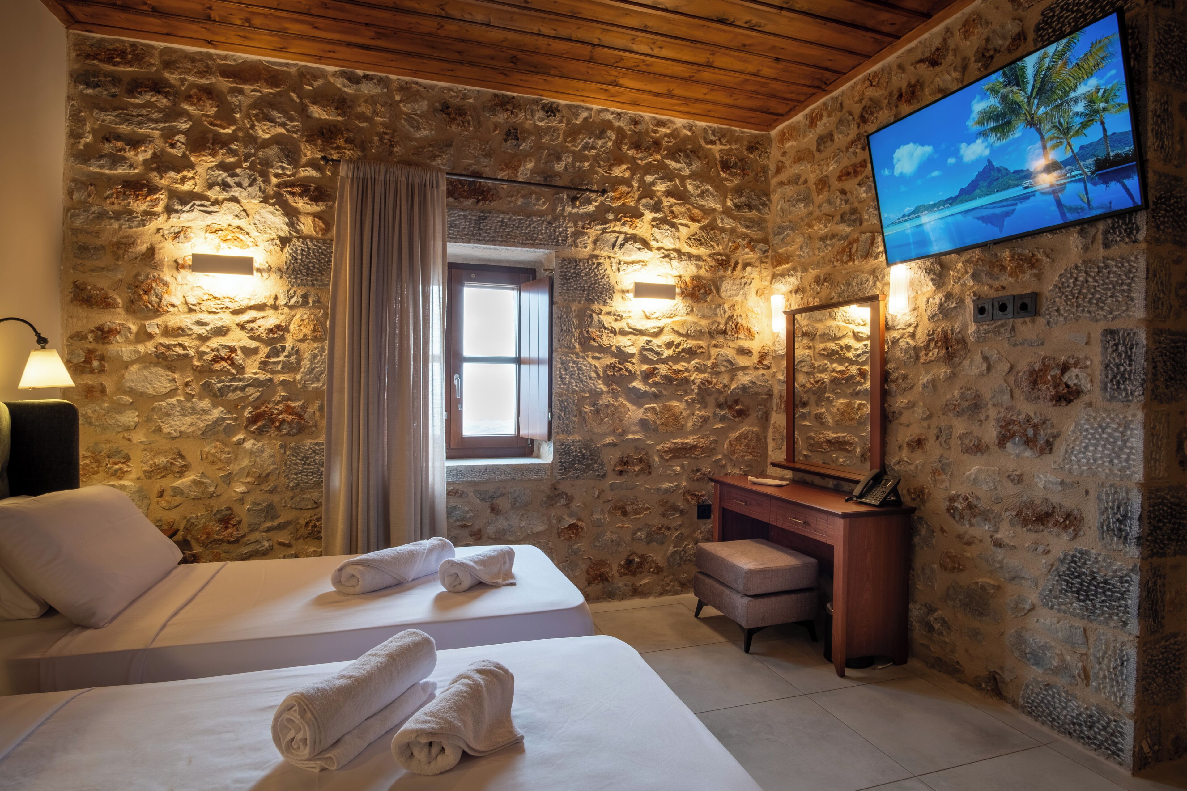 A room with stone walls, a wooden ceiling, a TV on the wall, a desk, and two beds with towels.