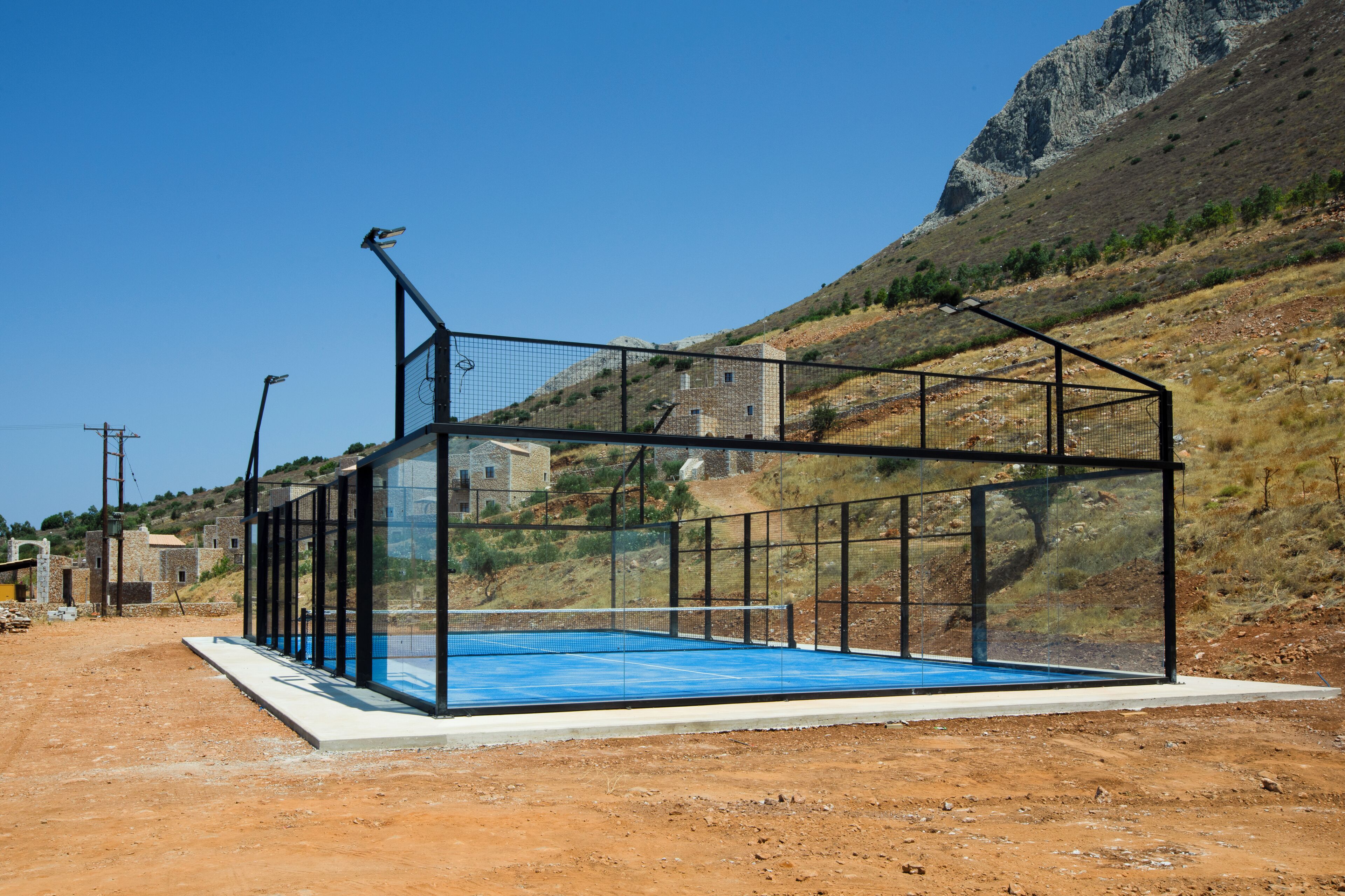 A tennis court with a net, surrounded by a metal fence, set in a hilly area with buildings in the background.