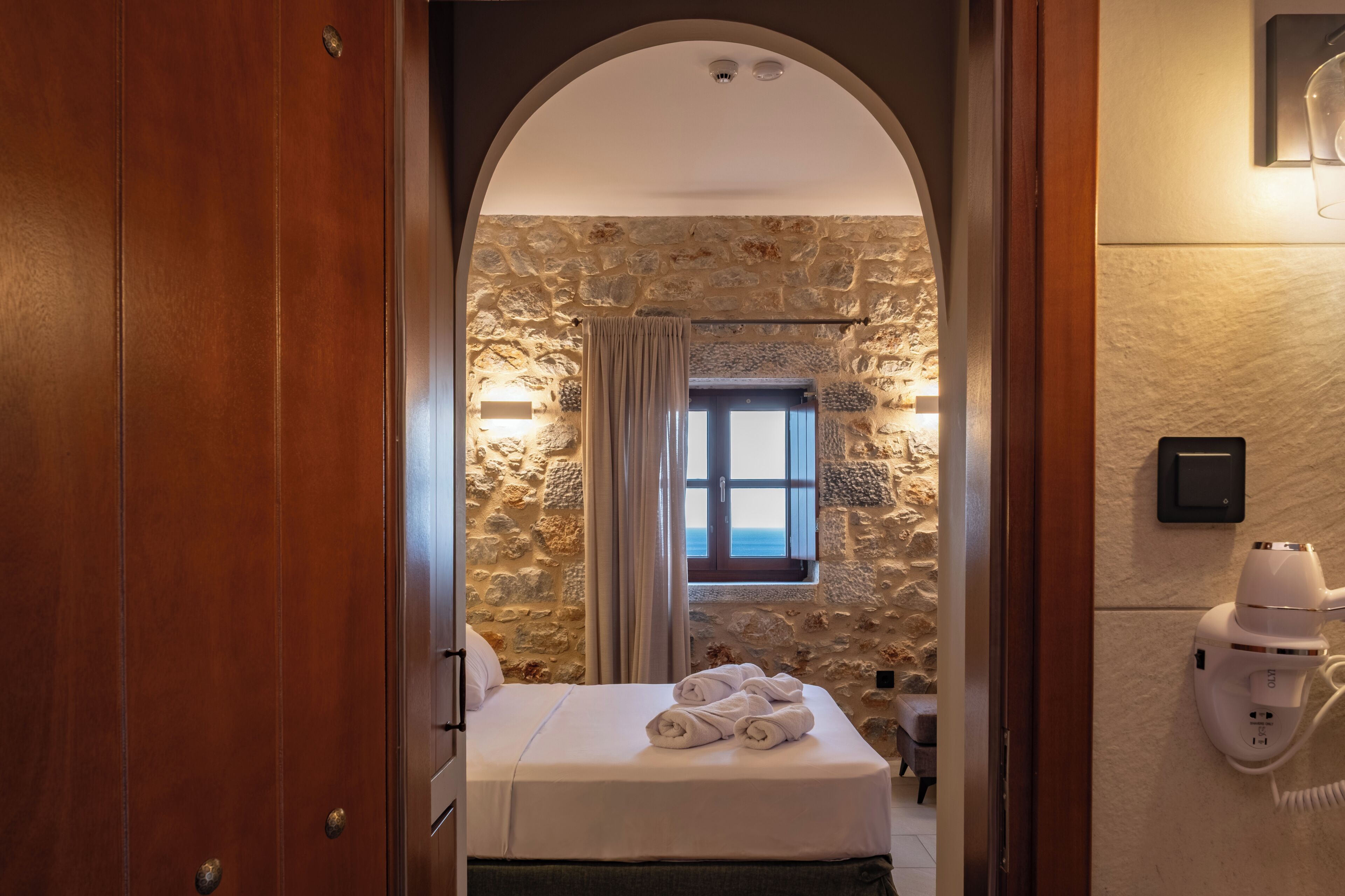 A hotel room with a stone wall, a bed with white linens, a window with curtains, and a hairdryer on the wall.