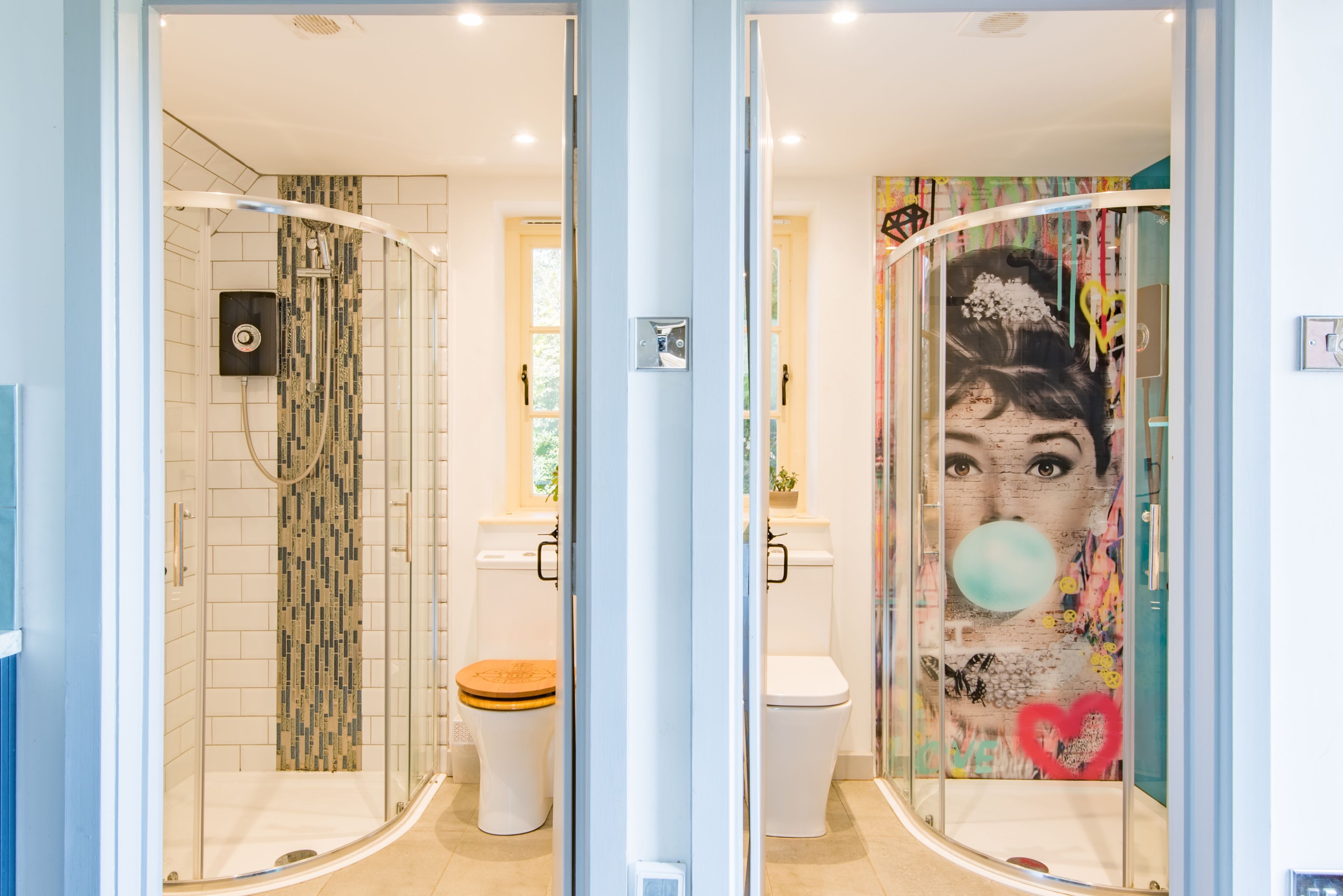 Une salle de bain avec une douche à parois de verre, des toilettes avec un abattant en bois et un mur orné d’une fresque colorée.