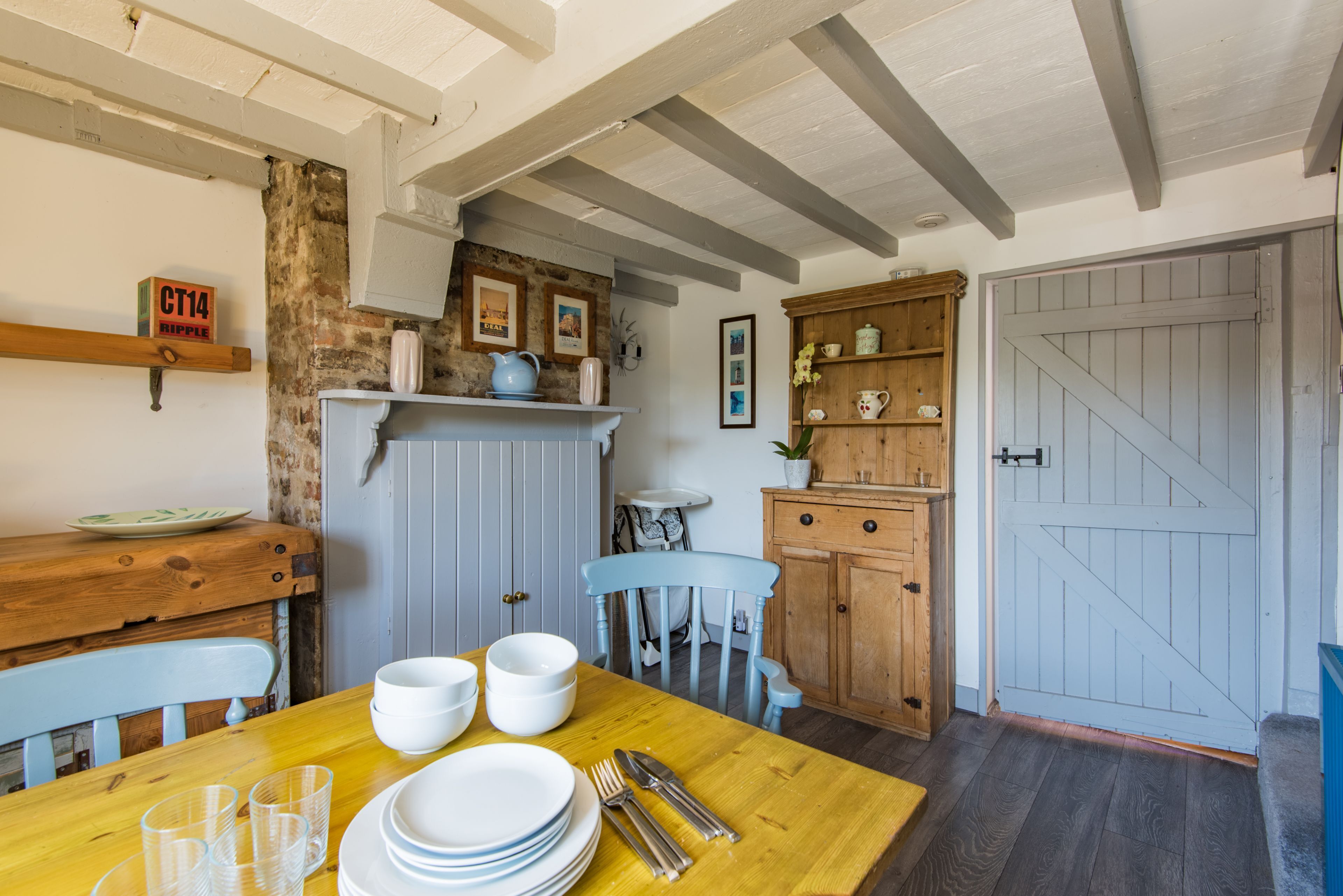 Un coin repas avec une table en bois dressée pour quatre personnes, une chaise bleue et, au fond, une cuisine au style rustique.