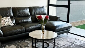 A modern living room with a black leather sofa, a round coffee table with a vase of flowers, and a patterned rug. - 2BR-2BA - Rivonia Residence 3 (Sandton)