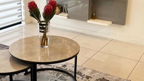 A modern living room with a flat-screen TV mounted on the wall, a round coffee table with a vase of flowers, and a patterned rug. - 2BR-2BA - Rivonia Residence 3 (Sandton)